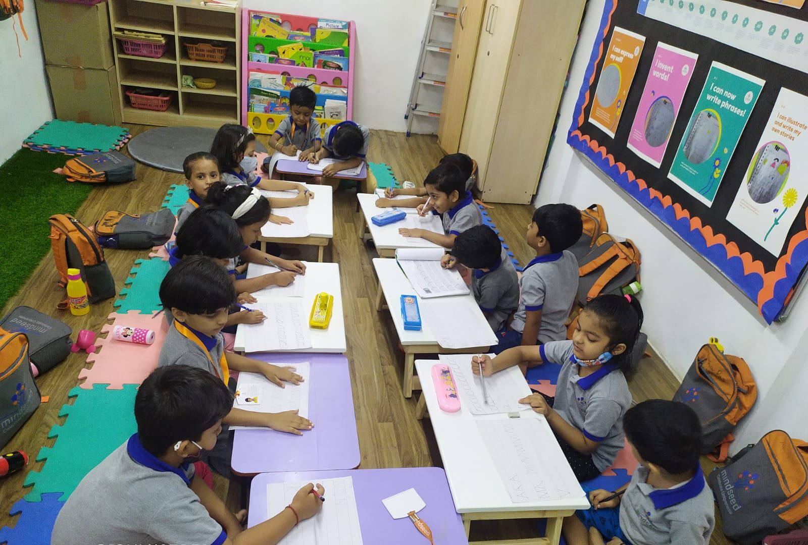 Mindseed kindergarten - A group of young students in matching uniforms sitting at small desks in a classroom, focused on writing or drawing in their notebooks.