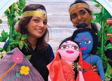 Two women wearing golden headbands pose with handmade puppets of Rama and Sita for a Mindseed preschool and daycare event.